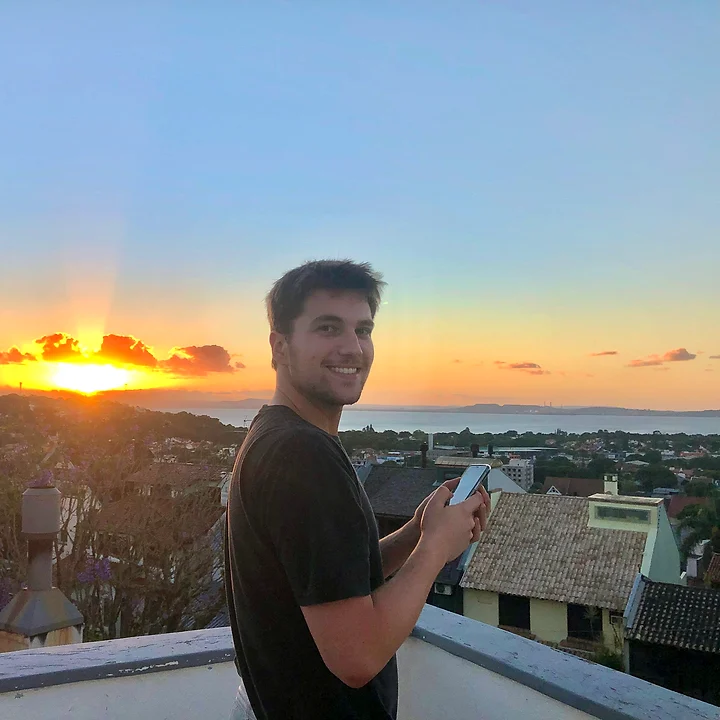 Bruno smiling on a rooftop at sunset, phone in hand, a city and the water behind him.